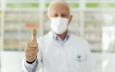 Focus on thumb up, male pharmacist showing a sign of approval with his hand. A man with gray hair in a white uniform and a protective face mask, gesturing approval with one hand. Look at the camera